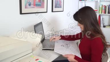 设计工程师坐在她办公室的办公桌前，在笔记本电脑上打字，在她的办公桌上放蓝图